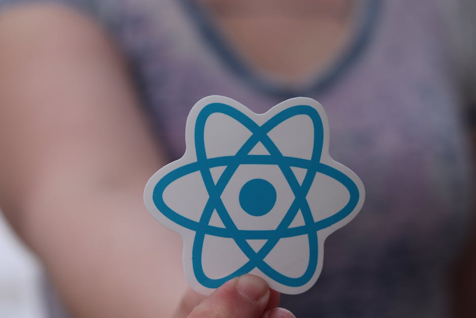 Close-up of a person's hand holding a sticker featuring a blue atom symbol with orbiting electrons, held in front of a blurred background that includes part of a person's arm and a indistinct indoor s