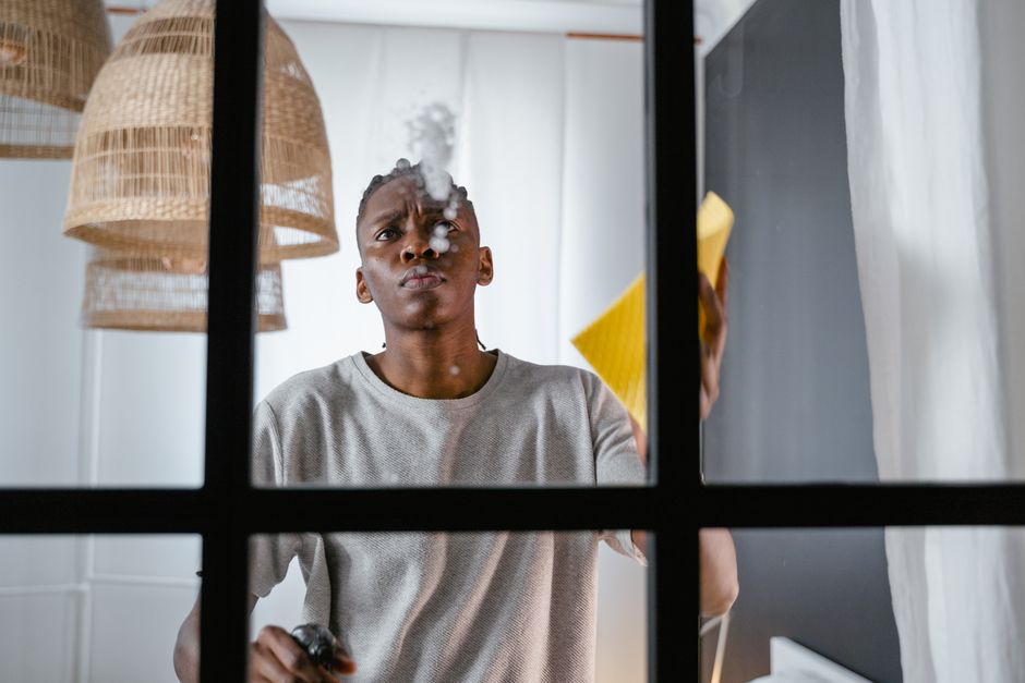 A young man with a serious expression is seen from inside a modern room, viewed through a partially open black-framed glass window. He is wearing a light gray T-shirt and is using a yellow cleaning cl