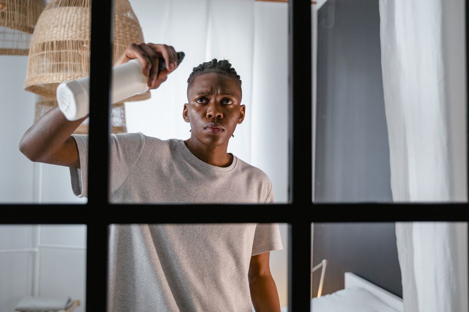 A young man with a serious expression is seen from inside a modern room, viewed through a partially open black-framed glass window. He is wearing a light gray T-shirt and is using a yellow cleaning cl